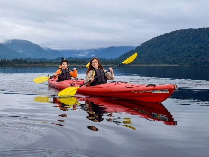 Two young woman smiling and kayaking on a mirror lake in Franz Josef, the South Island of New Zealand, during winter while on a ski and explore tour with Wild Kiwi.