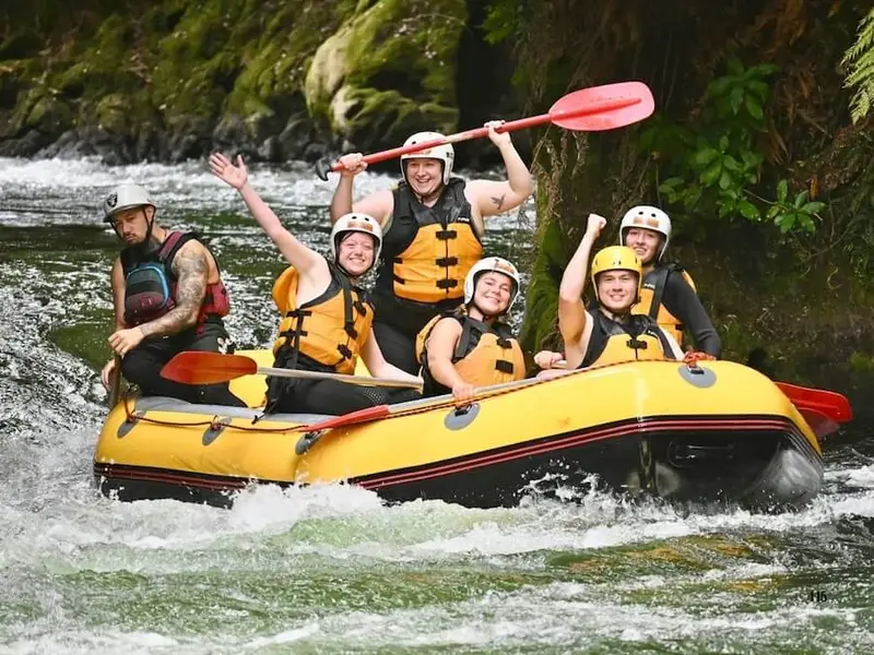 Group of travellers white-water rafting on the Kaituna River near Rotorua, New Zealand, smiling as they paddle through native forest before reaching Tutea Falls.