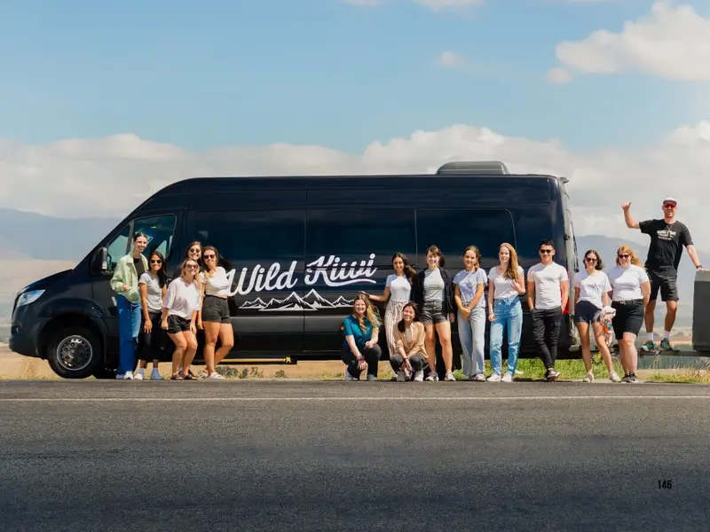 A Wild Kiwi small group tour standing beside a branded tour vehicle on a scenic New Zealand road trip, showing travellers exploring Aotearoa together by coach with open landscapes and mountains in the background, ideal for a North Island vs South Island New Zealand guide about choosing the best island, itinerary and small group adventure tour.