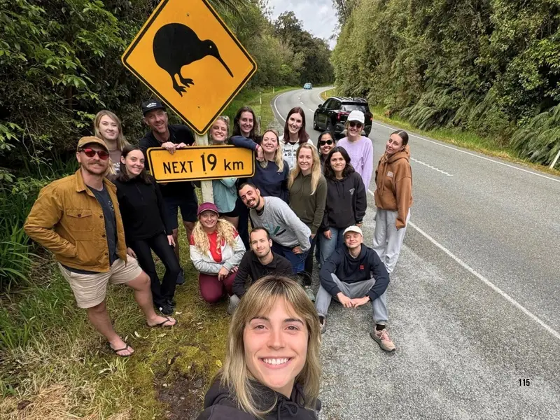 Travellers posing beside a Kiwi crossing road sign on a New Zealand road trip, surrounded by native forest.