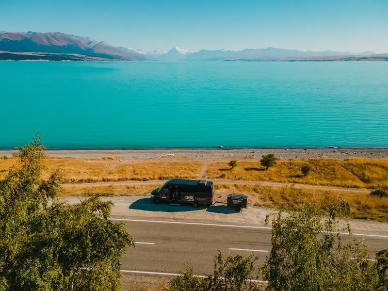 A Wild Kiwi tour vehicle parked beside the bright turquoise water of Lake Pukaki on New Zealand’s South Island, with open road views, golden grassland and the Southern Alps in the distance, showing the dramatic lake and mountain scenery that makes the South Island a top choice for road trips and small group adventure tours.