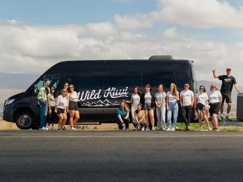 Wild Kiwi small group tour standing beside their black road trip van on a scenic New Zealand highway with mountains in the background.