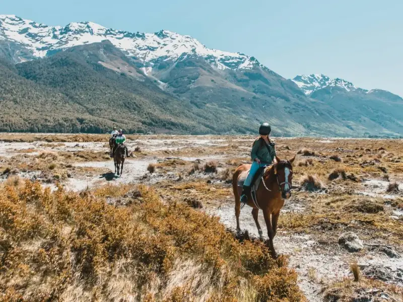 Travellers horse riding through an open South Island valley with snow-capped mountains in the distance, highlighting outdoor adventure, scenic landscapes and non-ski winter activities to add to a New Zealand travel itinerary.