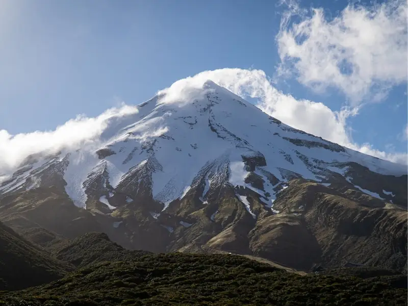 A close view of snow-covered Mount Taranaki with clouds around the summit and native bush below, highlighting one of New Zealand’s most iconic volcanic regions for North Island hikes and outdoor adventure travel.