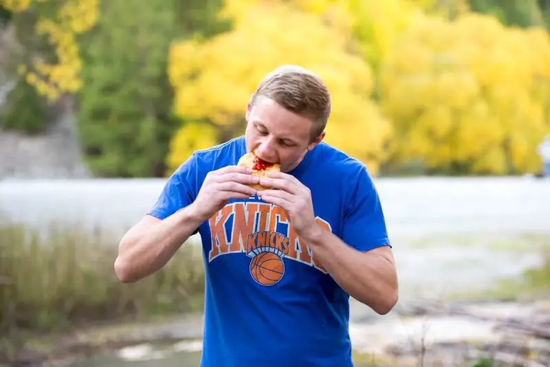 Traveller eating a classic New Zealand meat pie outdoors during a road trip, surrounded by autumn trees near a river.