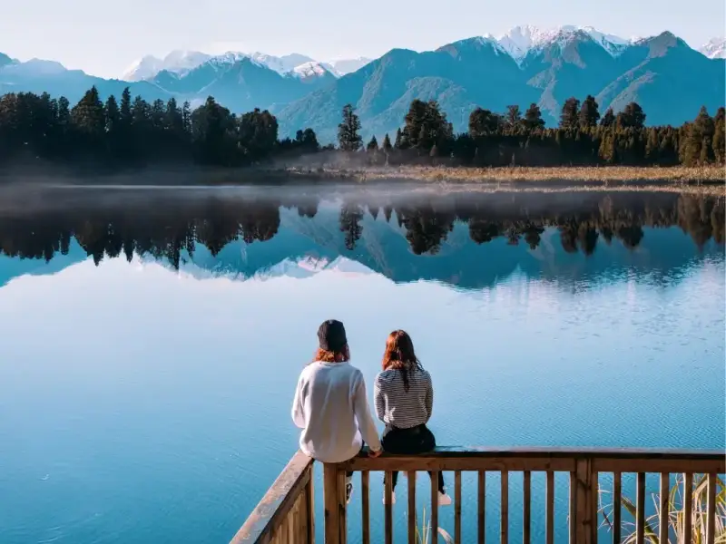 Travellers sitting beside a calm South Island lake with snow-capped mountains reflected in the water, highlighting peaceful New Zealand winter scenery, digital detox travel and scenic road trip stops beyond skiing.