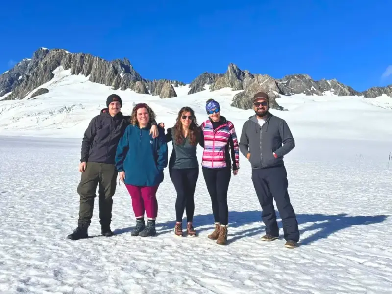 Travellers standing on a snowy glacier landscape with bright blue sky and alpine peaks behind them, showcasing a South Island winter adventure in New Zealand with glacier views, mountain scenery and small group travel experiences.