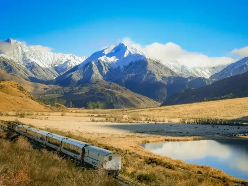 The TranzAlpine train winds through open alpine country beneath snow-capped peaks in New Zealand’s South Island. With reflective water, dry grassland and dramatic mountain scenery, this image highlights the striking contrasts of the TranzAlpine route as it crosses between Christchurch, Arthur’s Pass and Greymouth.