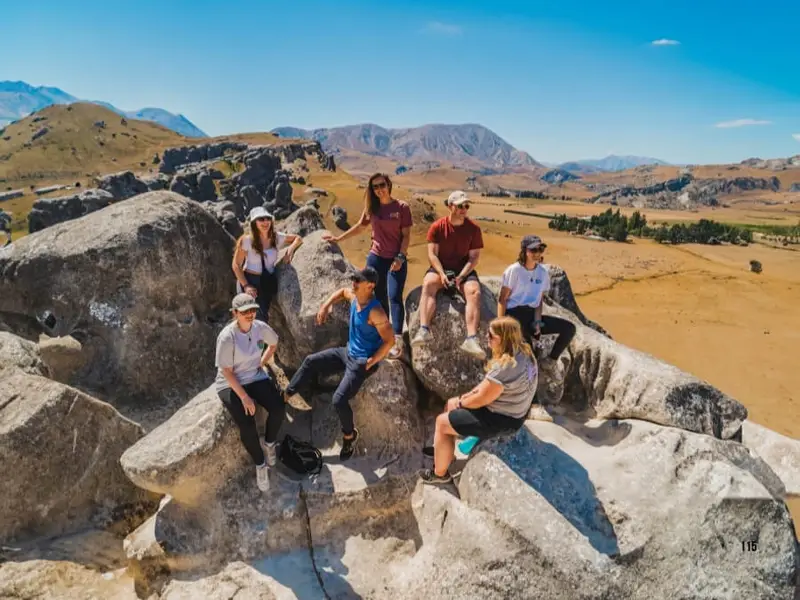 Travellers sitting on the giant limestone boulders at Castle Hill in New Zealand, enjoying sunny mountain views during an adventure tour.