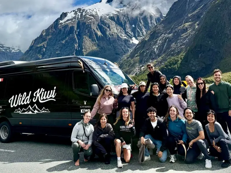 A Wild Kiwi small group tour posing beside a branded tour vehicle in front of dramatic South Island mountain scenery, highlighting the alpine landscapes, guided road trip experience and social adventure travel that make the South Island a top choice for travellers comparing North Island vs South Island New Zealand.