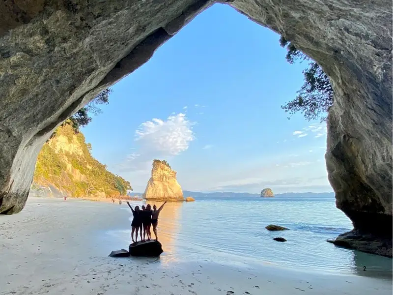 A group of travellers looking out from the Cathedral Cove rock arch toward the beach and ocean on the Coromandel Peninsula, a scenic North Island coastal walk featured in New Zealand hiking itineraries.