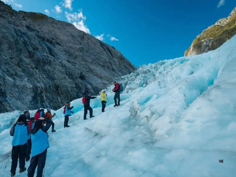 Travellers hiking across a bright blue glacier on New Zealand’s South Island, surrounded by rocky alpine scenery and mountain views, highlighting the dramatic glacier landscapes, guided outdoor adventure and bucket-list experiences that make the South Island a top choice for visitors comparing North Island vs South Island New Zealand.