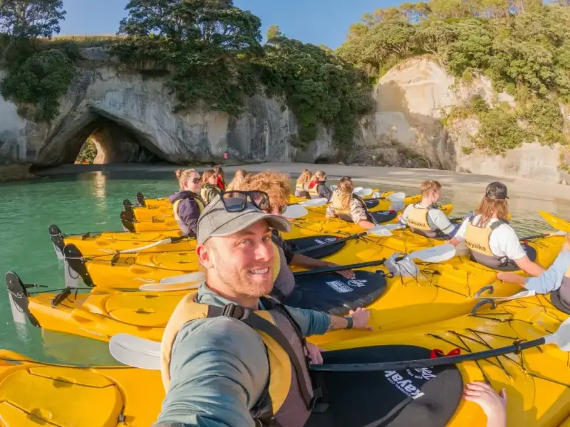 Group photo of young travellers in kayaks with the iconic Cathedral Cove of New Zealand behind them during a Srping tour in the North Island with Wild Kiwi.