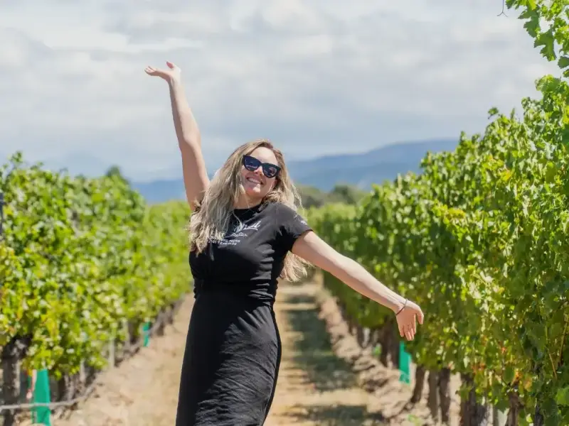 A smiling traveller stands between bright green vineyard rows in Marlborough, New Zealand, with arms outstretched on a sunny day in wine country. The photo reflects the relaxed and memorable experience of visiting Marlborough for Sauvignon Blanc tasting, vineyard tours, and scenic South Island travel, giving the article a more personal and experience-led feel.