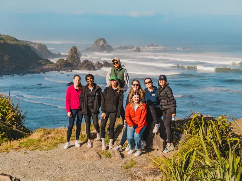 A group of female backpackers pose on cliffs with dramatic seascape behind on a sunny spring day in New Zealand while on a South Island small group tour.