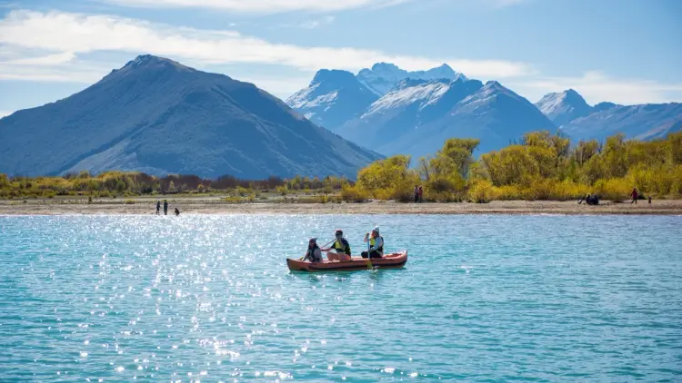 A wide autumn scene in New Zealand’s South Island showing travellers kayaking on a clear turquoise lake, with golden autumn trees along the shoreline and dramatic snow-dusted mountains in the background, capturing why March, April and May are ideal for New Zealand autumn travel, scenic road trips, and outdoor adventure.