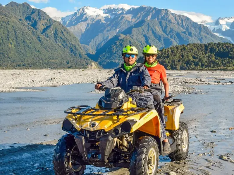 Two travellers riding a quad bike across a wide riverbed with rugged Southern Alps near Fox Glacier with mountains behind them, a high-energy New Zealand adventure activity that pairs perfectly with an autumn road trip itinerary.