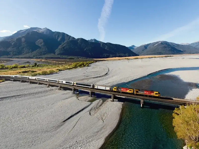 An aerial view of New Zealand’s TranzAlpine train crossing a long bridge over a braided river in the South Island, surrounded by wide gravel plains, clear blue water and rugged mountain ranges under a bright sky. This scenic TranzAlpine rail journey image captures the dramatic landscapes between Christchurch and Greymouth that make the route one of the most beautiful train journeys in New Zealand.