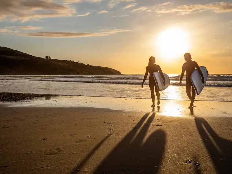 Two surfers walking along the beach at sunset in Raglan New Zealand, carrying surfboards with golden light reflecting off the water.