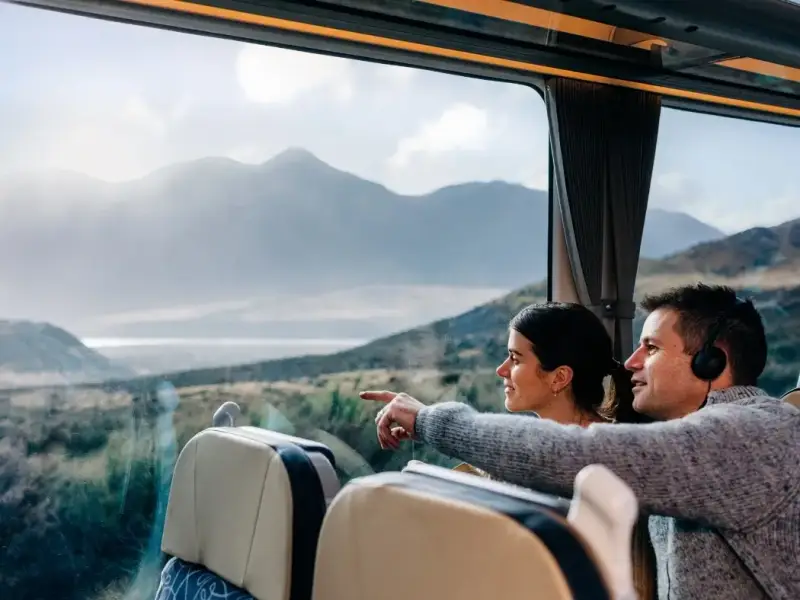 Passengers look out from the TranzAlpine train at sweeping South Island scenery in New Zealand, with mountains, open valleys and soft natural light beyond the carriage window. The image captures the relaxed onboard experience and scenic views that make the TranzAlpine train journey one of the best rail trips in New Zealand.