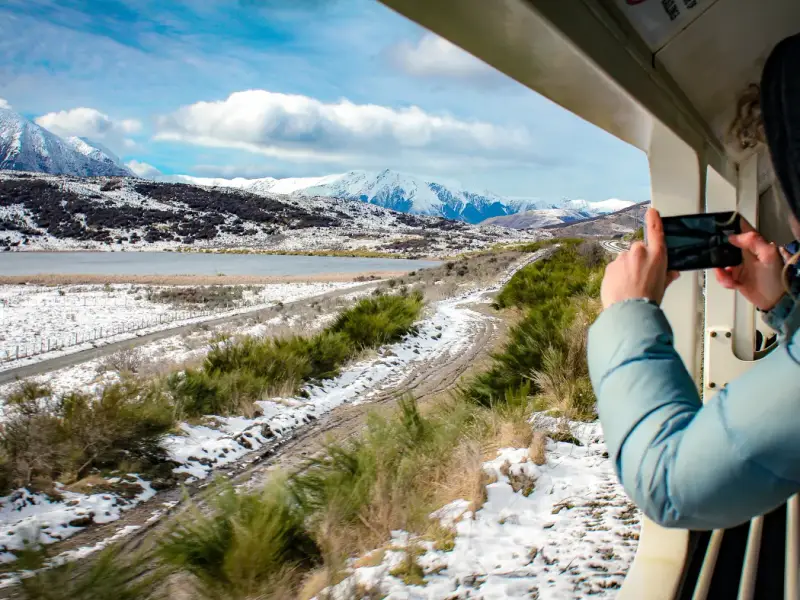 Traveller photographing snowy mountain scenery from a train window in New Zealand, showcasing a scenic winter rail journey with alpine landscapes, snow-covered valleys and a memorable way to experience South Island winter travel.