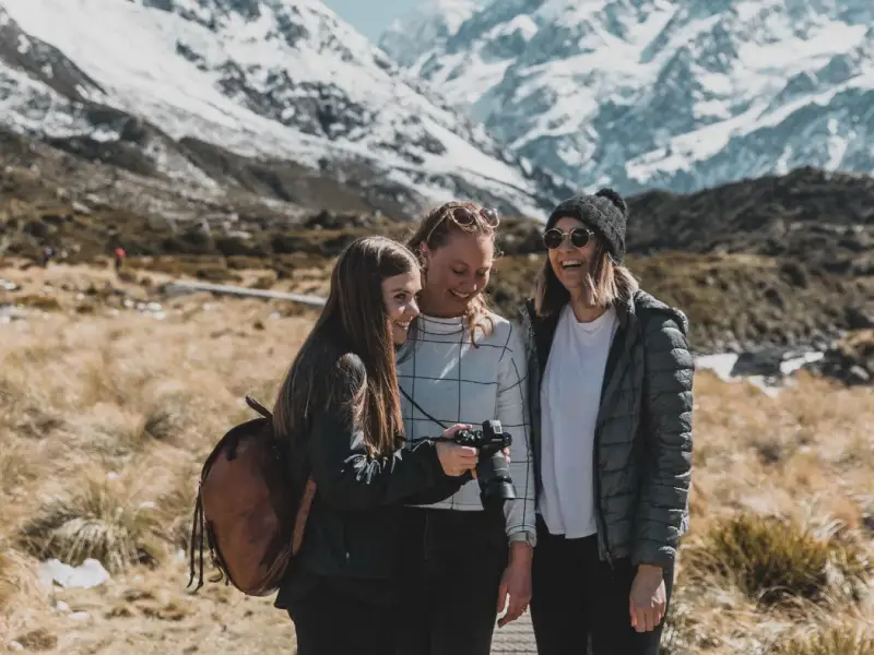 Travellers smiling with a camera in front of snow-capped South Island mountains, showing New Zealand winter travel beyond skiing with small group adventure, alpine scenery, photography stops and scenic outdoor experiences.