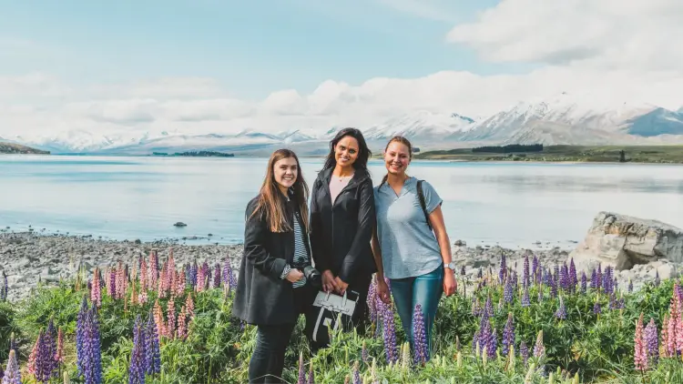 Three women travellers stand by a bright blue lake surrounded by lupins and snow-capped mountains in New Zealand, capturing the spirit of female travel, friendship, and support on a Wild Kiwi road trip. This image suits content about periods while travelling, solo female travel in New Zealand, and how Wild Kiwi helps women feel comfortable and looked after on the road.