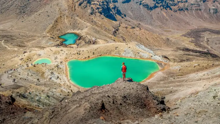A hiker standing above the Emerald Lakes on the Tongariro Alpine Crossing, with volcanic terrain and turquoise crater lakes across Tongariro National Park, one of the most iconic New Zealand hiking trails.
