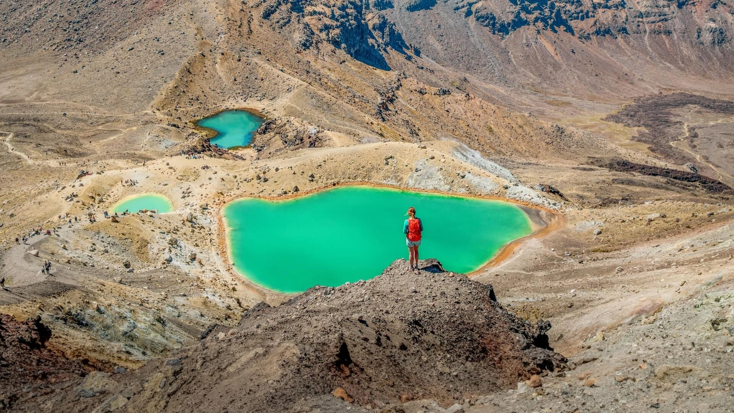 A hiker standing above the Emerald Lakes on the Tongariro Alpine Crossing, with volcanic terrain and turquoise crater lakes across Tongariro National Park, one of the most iconic New Zealand hiking trails.