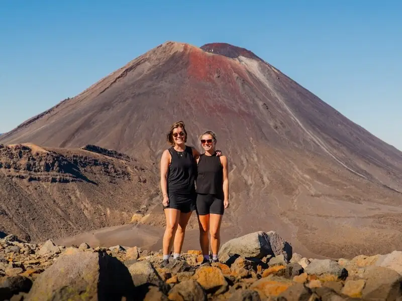 Two hikers standing in front of Mount Ngauruhoe on the Tongariro Alpine Crossing, New Zealand, under clear blue skies.