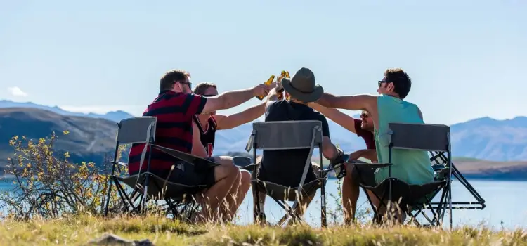 A group of people cheers drinks in a field near a lake with a backdrop of mountains in New Zealand.