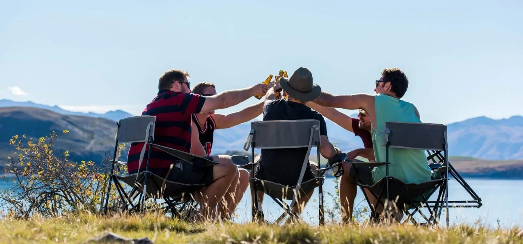 A group of people cheers drinks in a field near a lake with a backdrop of mountains in New Zealand.