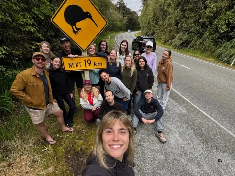 Wild Kiwi travellers on a New Zealand group tour taking a roadside selfie next to a large kiwi bird warning sign and “NEXT 19 km” sign on a forest-lined highway.