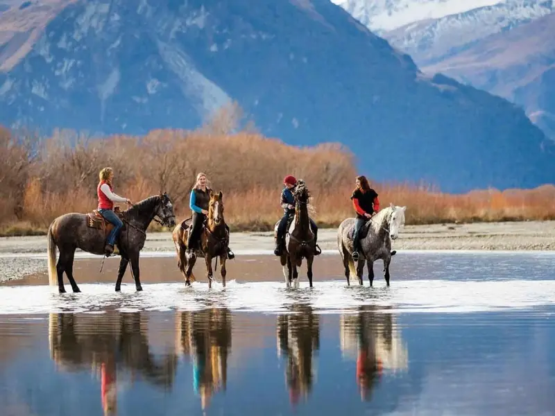 Group horse trekking through shallow river water with mountains rising behind them, a memorable New Zealand adventure experience that works especially well in autumn with cooler days and quieter trails.