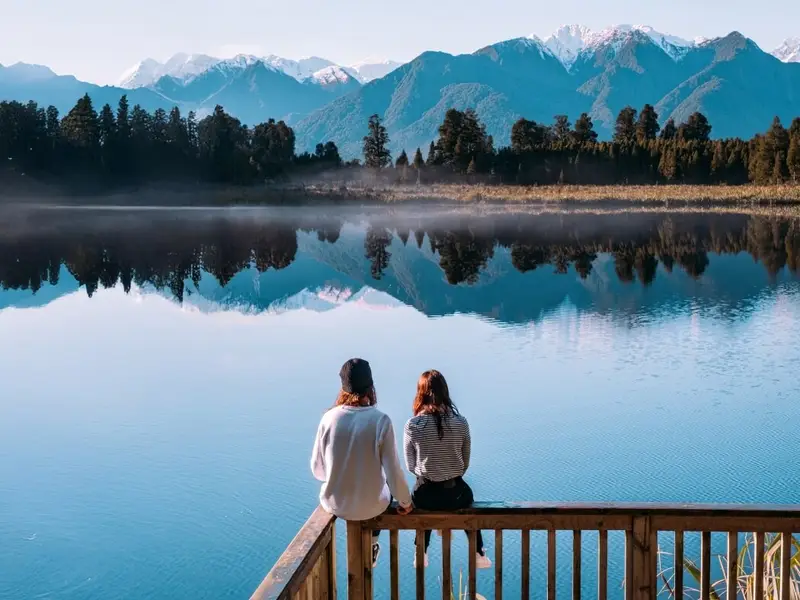 Two cyclists riding beside a bright turquoise lake with golden autumn shrubs and wide mountain views, a perfect example of why autumn is one of the best times to visit New Zealand for active days outdoors.