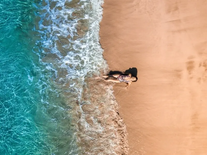 Traveller relaxing at the edge of turquoise waves on a golden New Zealand beach, captured by drone during summer.