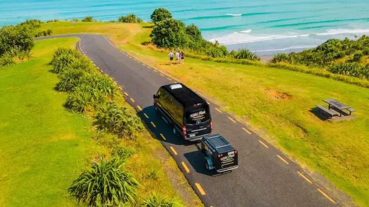 Wild Kiwi van driving along a coastal New Zealand road near Raglan with turquoise ocean waves and travellers enjoying the view — promoting the Wild Kiwi Northern Voyager trip giveaway.