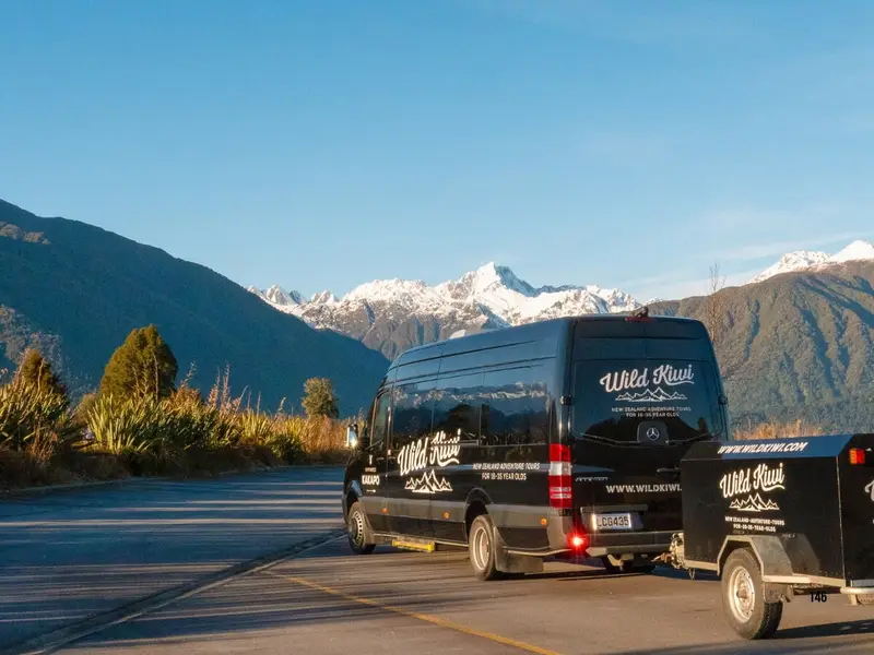 A Wild Kiwi tour vehicle driving through dramatic South Island mountain scenery in New Zealand, with snow-capped peaks and open road views in the background, highlighting the guided road trip experience, alpine landscapes and adventure travel style that make the South Island a top choice for travellers comparing North Island vs South Island New Zealand.
