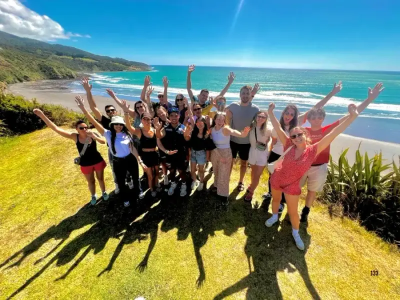 Group of Wild Kiwi travellers on a New Zealand adventure tour with arms raised at a scenic coastal viewpoint overlooking the ocean and beach shoreline.