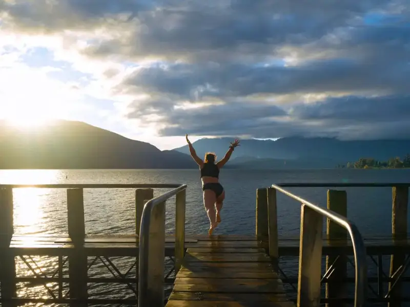 Traveller jumping from a wooden jetty into a New Zealand lake at sunrise, highlighting cold plunges, winter wellness and digital detox travel as refreshing activities to include in a New Zealand winter itinerary beyond skiing.