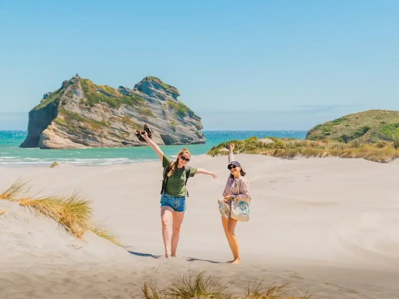 Two travellers exploring Wharariki Beach near Golden Bay, New Zealand, walking through sand dunes with the famous Archway Islands in the background on a sunny day.