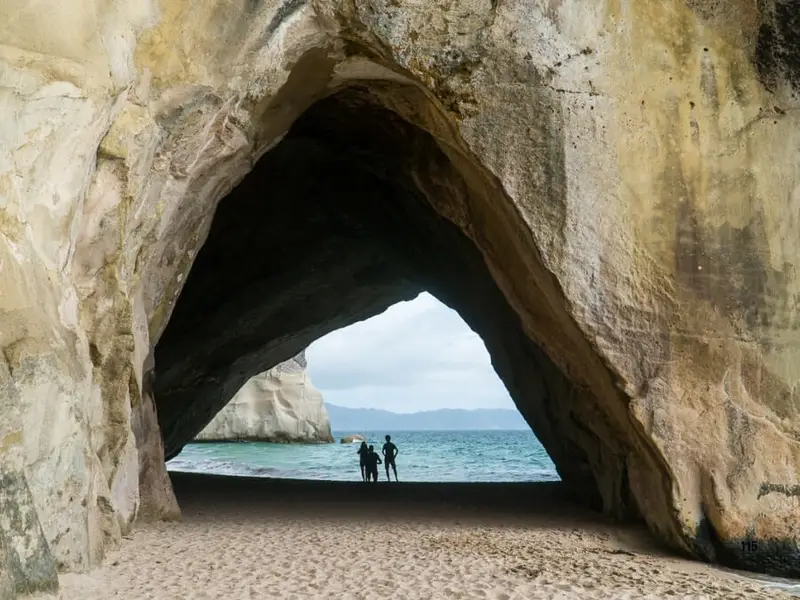 Silhouette of travellers standing inside the Cathedral Cove rock arch on the Coromandel Peninsula, looking out toward the turquoise sea.