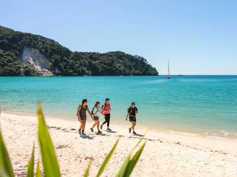 Group of travellers hiking the Abel Tasman Coast Track beside bright turquoise water and white sand beach, with native forest and anchored sailboats in the background.