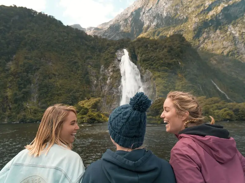 Friends laughing at a Milford Sound viewpoint with a tall waterfall cascading in the background, capturing the social side of New Zealand travel and the cooler, calmer feel of visiting in autumn.