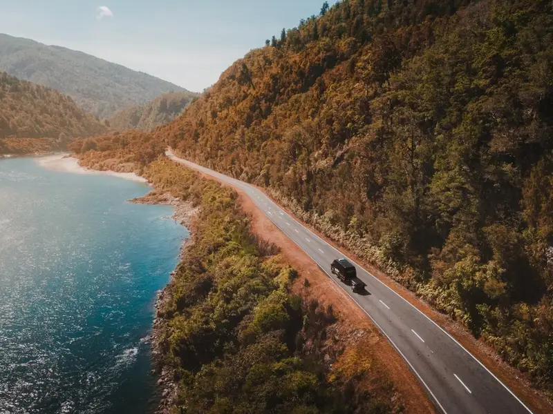 Aerial view of a winding coastal road with a vehicle travelling beside blue water and forested hills, the kind of scenic drive that makes a New Zealand autumn road trip feel relaxed and unrushed.
