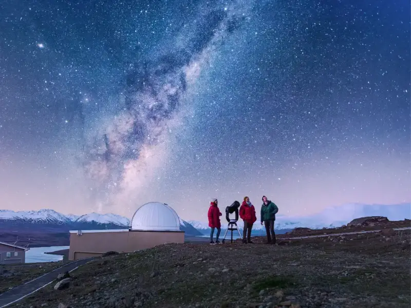 Travellers stargazing under the Milky Way near Lake Tekapo in New Zealand’s South Island, with an observatory, telescope, snow-capped mountains and clear winter night sky highlighting one of the best winter activities in New Zealand beyond skiing.