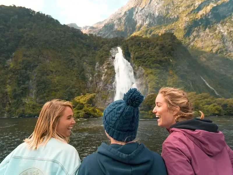 Smiling and happy solo female travellers on a Milford Sound day tour looking out at the raging waterfalls.