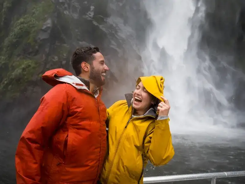 A couple laughs in front of a powerful waterfall while on Milford Sound day tour from Queenstown.