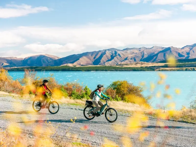 Two cyclists riding beside a bright turquoise Tekapo Lake with golden autumn shrubs and wide mountain views, a perfect example of why autumn is one of the best times to visit New Zealand for active days outdoors.