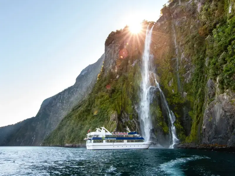 Scenic cruise boat passing beneath a waterfall in Milford Sound, showing one of New Zealand’s most iconic winter travel experiences with dramatic fjord cliffs, misty mountain scenery and powerful waterfalls on the South Island.